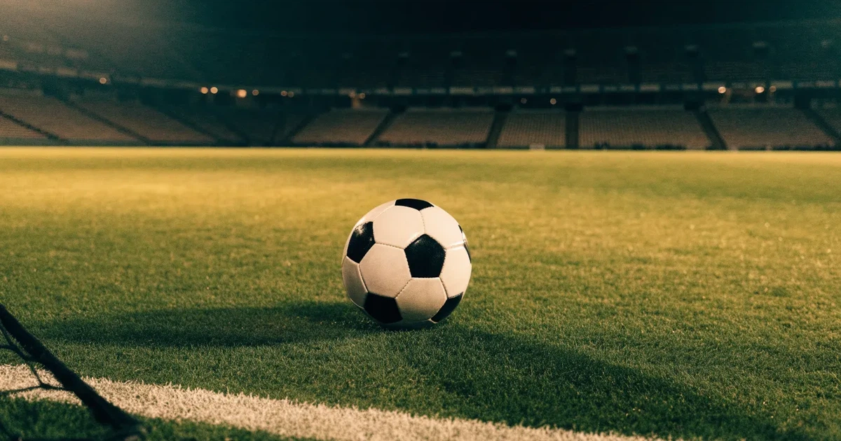 Ein Fußball liegt auf dem Rasen eines leeren Stadions bei Flutlicht, im Hintergrund unscharfe Tribünen ohne erkennbare Beschriftung.