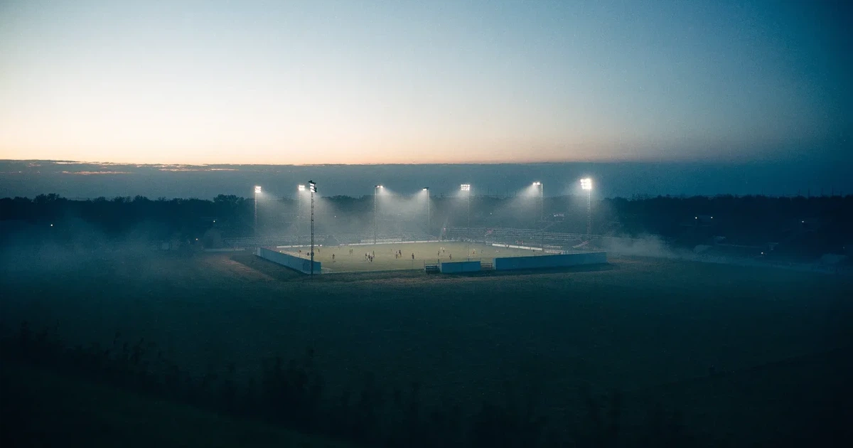 Ein Stadion unter Flutlicht, aus großer Entfernung über eine dunkle Wiese fotografiert, am Himmel zieht Nebel auf.