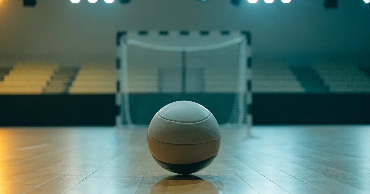 Ein Handball liegt auf dem Hallenboden vor einem Tor mit Netz, die leere Halle wird von Flutlicht beleuchtet.