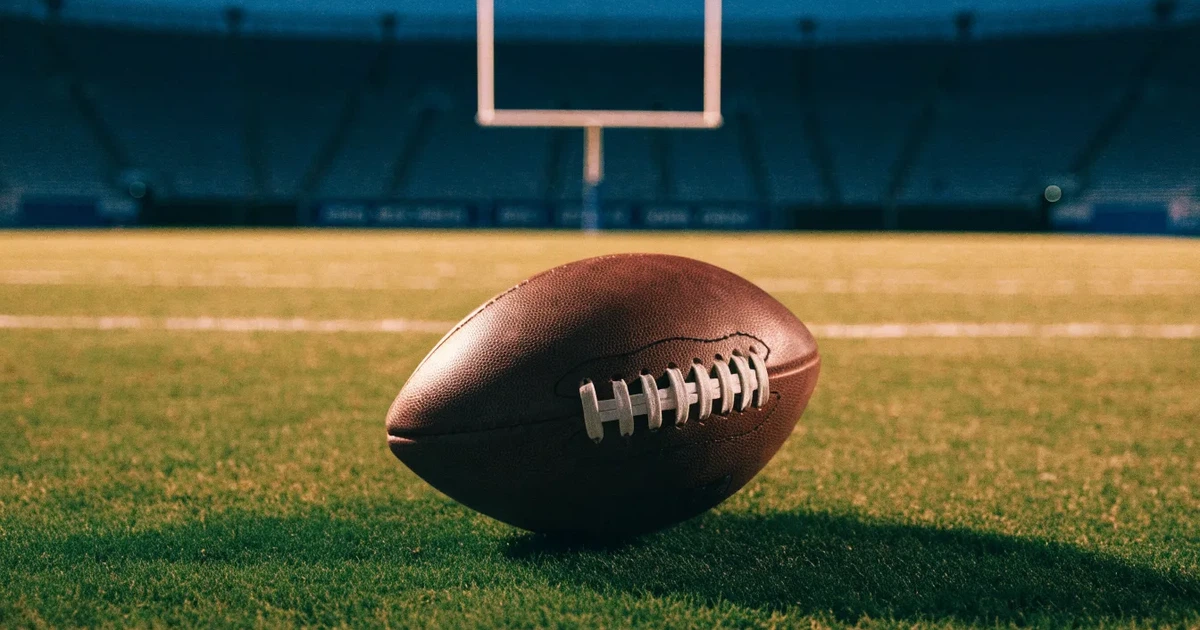 Ein American Football liegt auf dem Rasen eines leeren Stadions unter Flutlicht, die Torstangen ragen im Hintergrund auf.