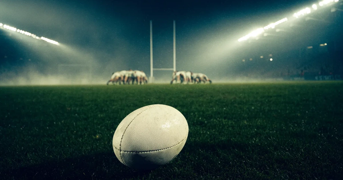 Ein ovaler Rugbyball liegt vor den hohen Torstangen auf nassem Rasen, über dem Stadion zieht Nebel auf.