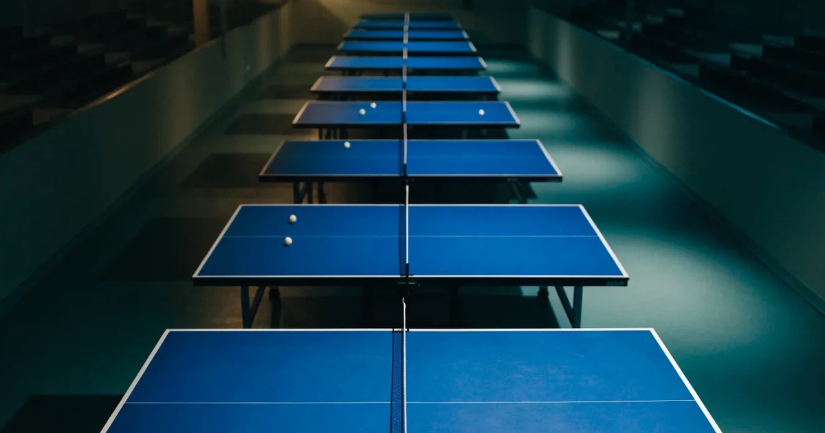Eine Tischtennishalle aus erhöhter Perspektive mit mehreren blauen Tischen in Reihen unter gedämpftem Hallenlicht, ohne Personen.