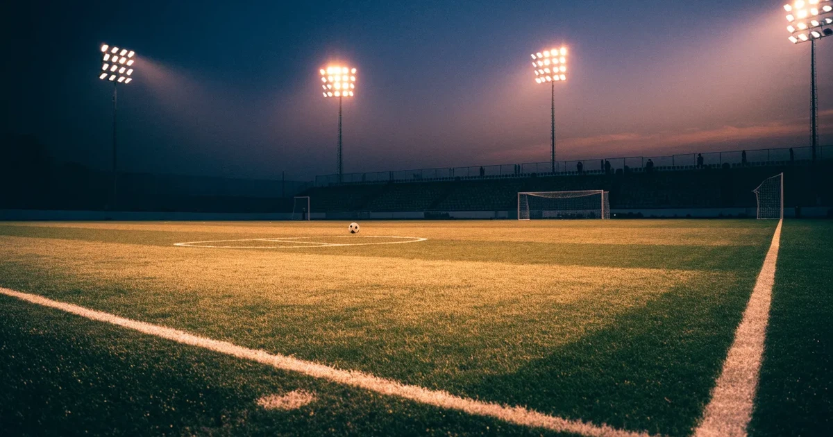 Ein Rasenplatz in der Dämmerung mit eingeschalteten Flutlichtmasten, ein Ball liegt am Anstoßpunkt, die Tribünen im Hintergrund sind unscharf.