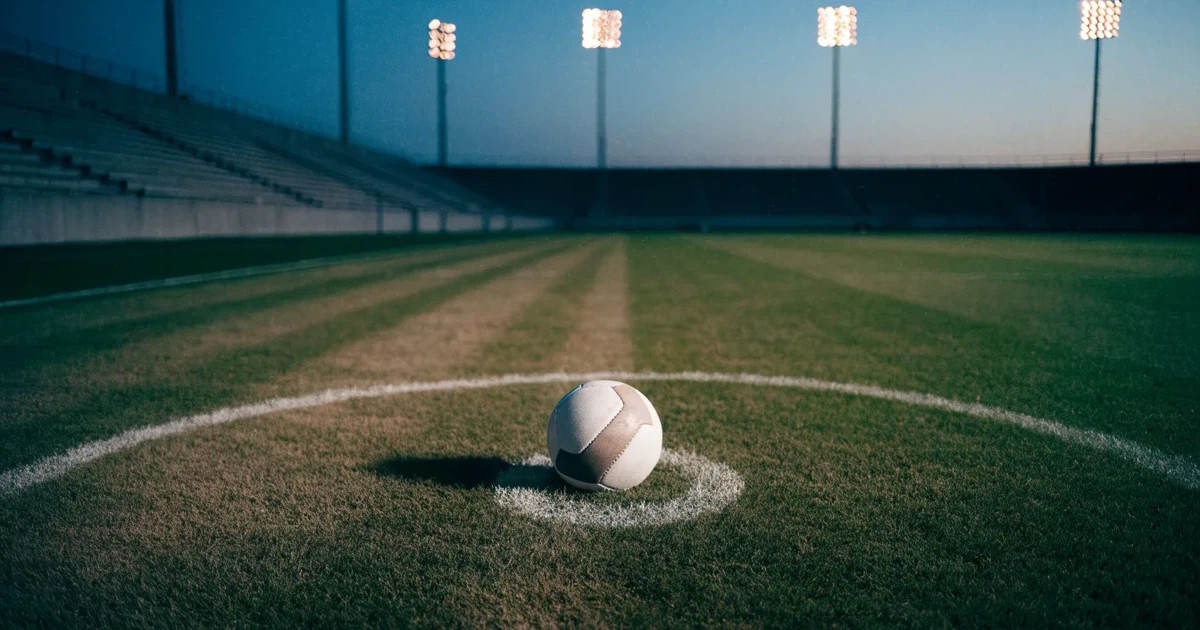 Ein Fußball liegt auf dem Anstoßpunkt eines leeren Rasenplatzes, im Hintergrund eine Tribüne und Flutlichtmasten in der Abenddämmerung.