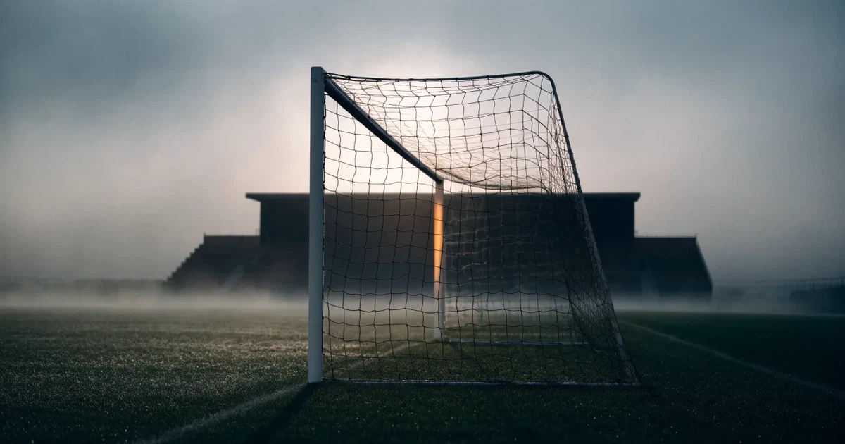 Ein Fußballtor mit Netz steht im Morgennebel auf einem leeren Rasenplatz, im Hintergrund die Silhouette einer kleinen Tribüne.