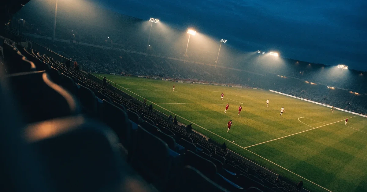 Ein Fußballstadion am Abend unter Flutlicht aus der Ferne gesehen, die Tribünen sind leicht beleuchtet und der Himmel dunkelblau.
