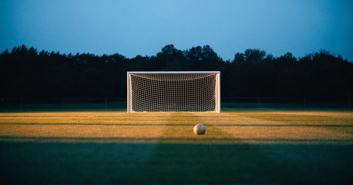 Ein Fußballtor mit Netz im Gegenlicht auf einem leeren Platz am Abend, davor ein Ball auf dem Rasen.